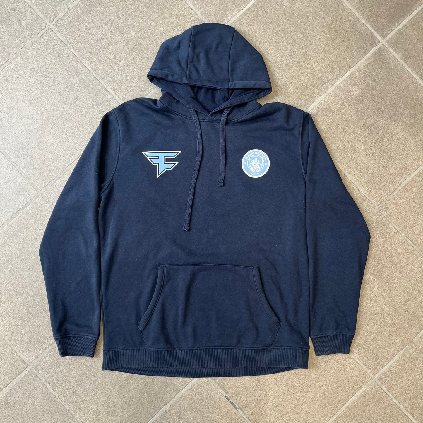 FaZe Clan x Manchester City 2019 Hoodie is an official collaboration combining esports and football culture. Made in Portugal from a 60% cotton and 40% polyester blend, it features the Manchester City club crest on the right chest and the FaZe Clan logo on the left. The back is finished with a large "FAZE CITY" graphic, with ribbed cuffs, a ribbed hem, and a kangaroo pocket.