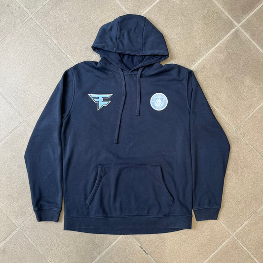 FaZe Clan x Manchester City 2019 Hoodie is an official collaboration combining esports and football culture. Made in Portugal from a 60% cotton and 40% polyester blend, it features the Manchester City club crest on the right chest and the FaZe Clan logo on the left. The back is finished with a large "FAZE CITY" graphic, with ribbed cuffs, a ribbed hem, and a kangaroo pocket.