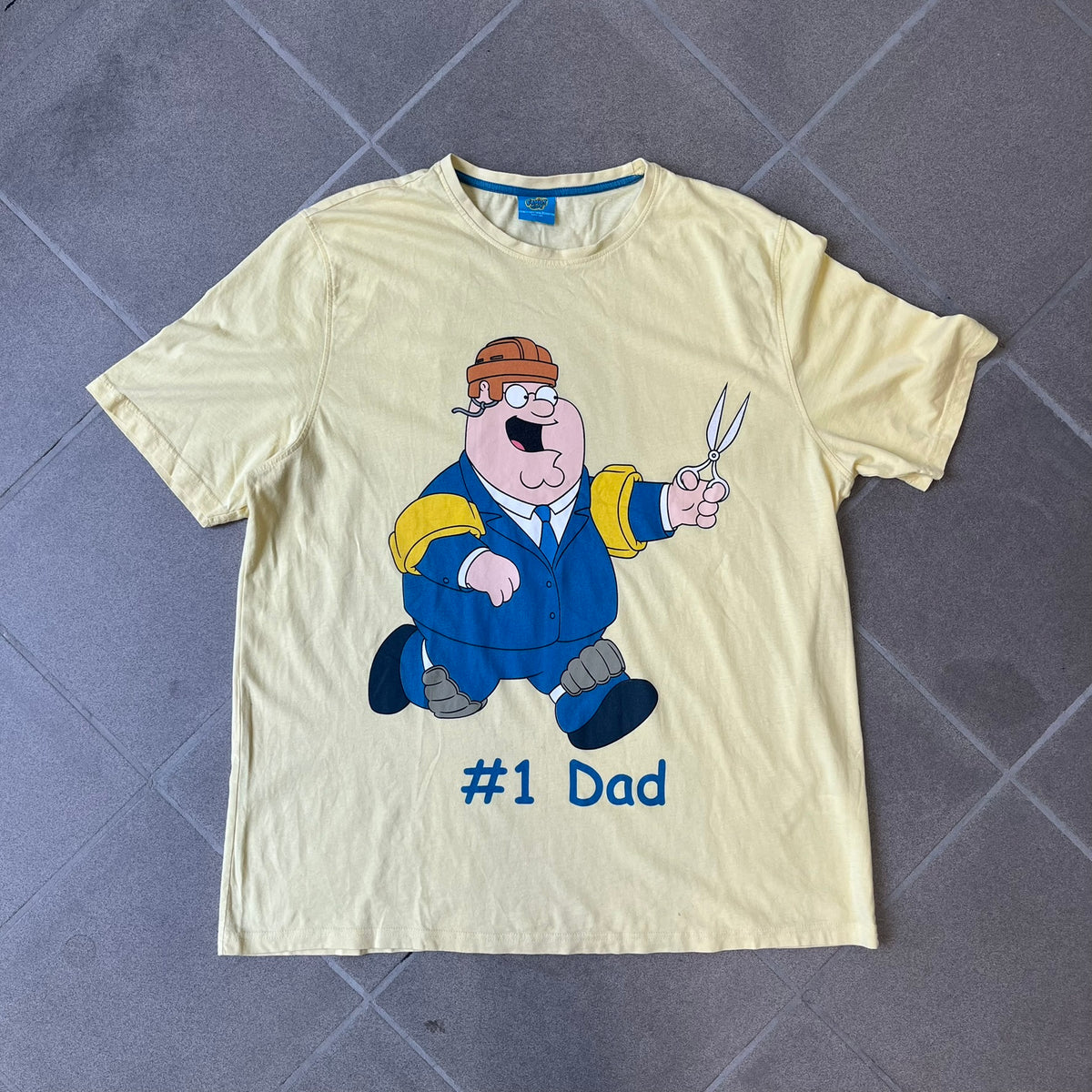 Family Guy 2012 Peter Griffin #1 Dad Tee features a yellow T-shirt with a front graphic of Peter Griffin wearing a helmet and elbow and knee protectors while running with a pair of scissors. A “#1 Dad” text is printed underneath the graphic. Includes a 2012 licensed neck tag.
