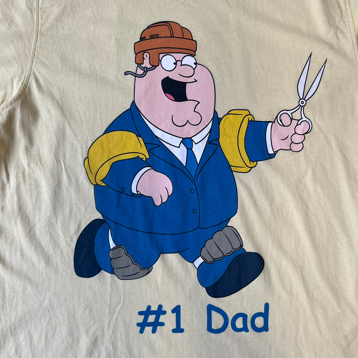 Family Guy 2012 Peter Griffin #1 Dad Tee features a yellow T-shirt with a front graphic of Peter Griffin wearing a helmet and elbow and knee protectors while running with a pair of scissors. A “#1 Dad” text is printed underneath the graphic. Includes a 2012 licensed neck tag.
