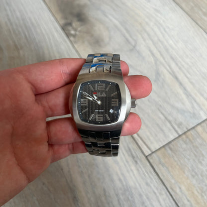 Fila Gents Quartz 320-G Analog Watch is a stainless steel wristwatch featuring a thick silver-tone case and bracelet. The dial has a black carbon-style texture with silver-tone numerals, hands, and a date display window. Powered by a Japanese Movt-2115 quartz movement, the watch includes a single crown and is rated for 10 ATM water resistance. Made in Japan.
