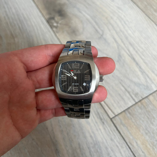 Fila Gents Quartz 320-G Analog Watch is a stainless steel wristwatch featuring a thick silver-tone case and bracelet. The dial has a black carbon-style texture with silver-tone numerals, hands, and a date display window. Powered by a Japanese Movt-2115 quartz movement, the watch includes a single crown and is rated for 10 ATM water resistance. Made in Japan.
