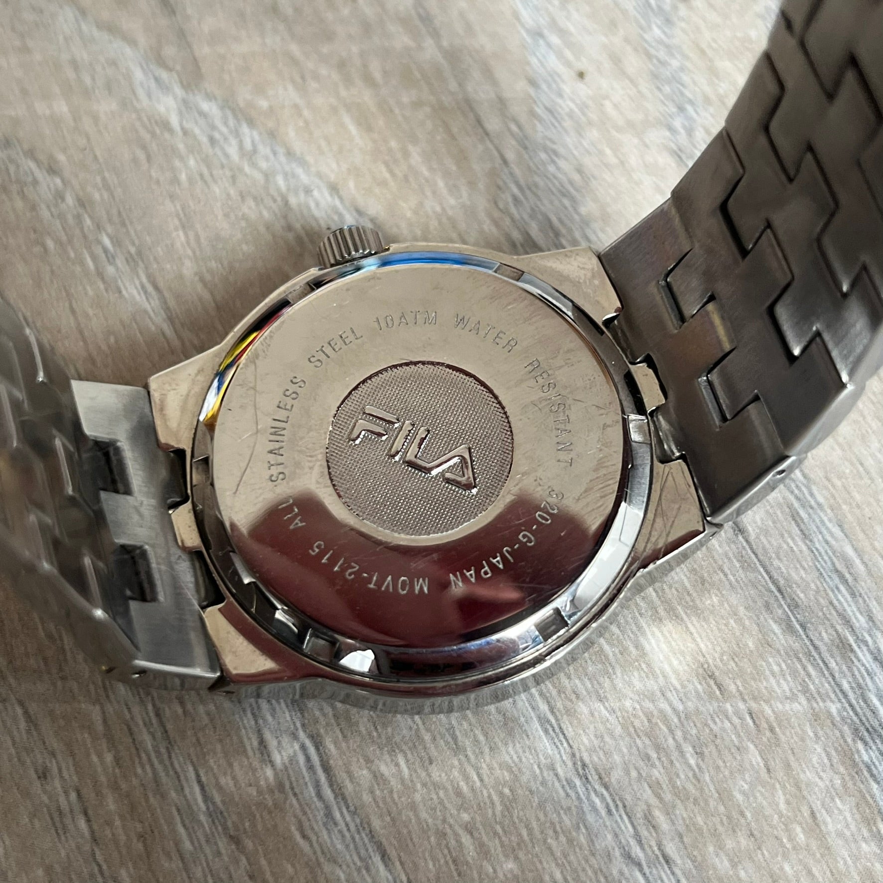 Fila Gents Quartz 320-G Analog Watch is a stainless steel wristwatch featuring a thick silver-tone case and bracelet. The dial has a black carbon-style texture with silver-tone numerals, hands, and a date display window. Powered by a Japanese Movt-2115 quartz movement, the watch includes a single crown and is rated for 10 ATM water resistance. Made in Japan.
