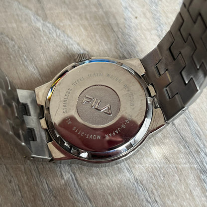 Fila Gents Quartz 320-G Analog Watch is a stainless steel wristwatch featuring a thick silver-tone case and bracelet. The dial has a black carbon-style texture with silver-tone numerals, hands, and a date display window. Powered by a Japanese Movt-2115 quartz movement, the watch includes a single crown and is rated for 10 ATM water resistance. Made in Japan.

