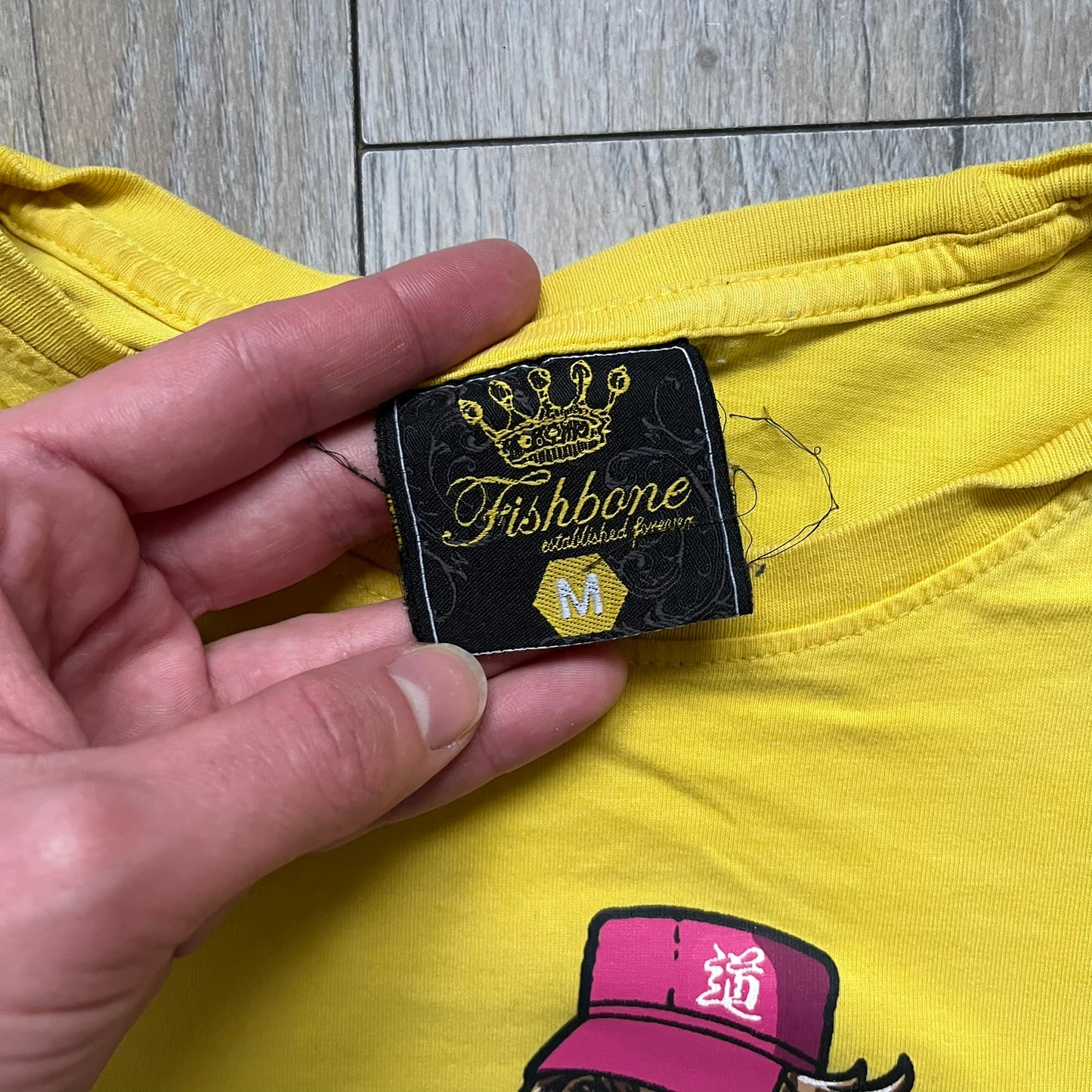 Fishbone Anime Girl 2000’s Yellow Tee is a yellow T-shirt featuring a large blue circle on the front with an anime-style naked girl wearing a pink hat and shorts. Made in the 2000s, it includes a black Fishbone neck tag.