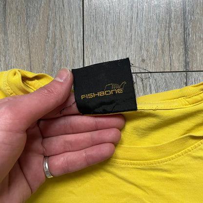Fishbone Anime Girl 2000’s Yellow Tee is a yellow T-shirt featuring a large blue circle on the front with an anime-style naked girl wearing a pink hat and shorts. Made in the 2000s, it includes a black Fishbone neck tag.