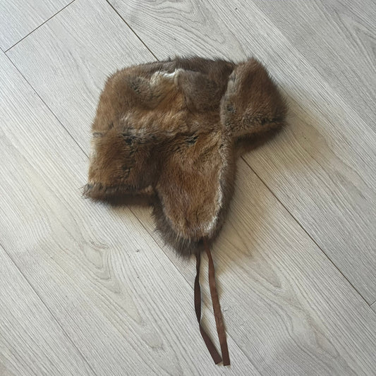 Fur ushanka hat featuring a Kulo tag inside and two leather tie strings. The front flap can be secured or folded back using small metal hooks for adjustable coverage. Designed for warmth and comfort, this classic winter piece offers both functionality and timeless style.