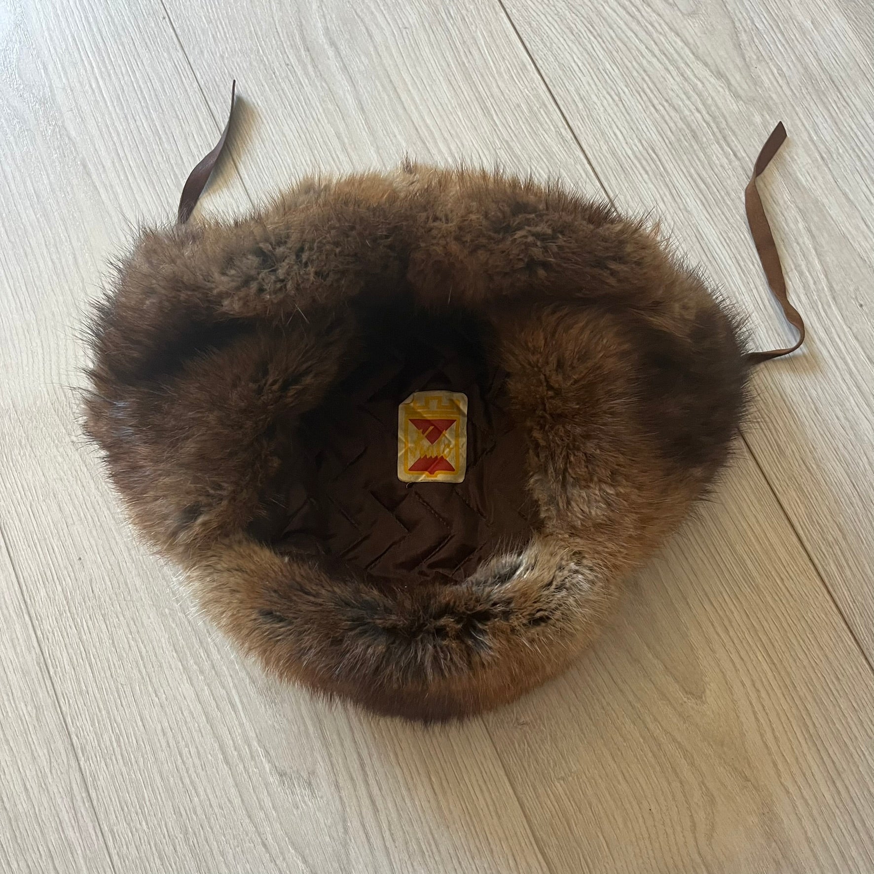 Fur ushanka hat featuring a Kulo tag inside and two leather tie strings. The front flap can be secured or folded back using small metal hooks for adjustable coverage. Designed for warmth and comfort, this classic winter piece offers both functionality and timeless style.