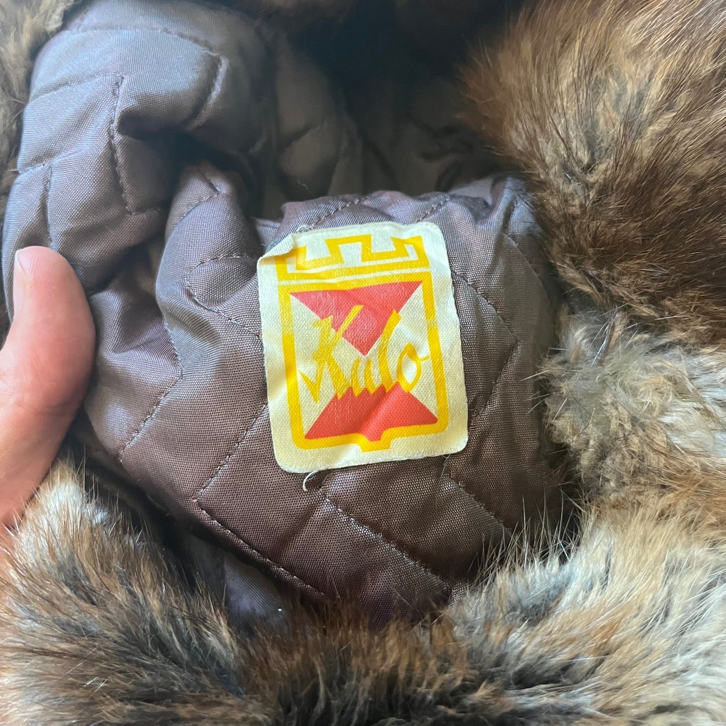 Fur ushanka hat featuring a Kulo tag inside and two leather tie strings. The front flap can be secured or folded back using small metal hooks for adjustable coverage. Designed for warmth and comfort, this classic winter piece offers both functionality and timeless style.