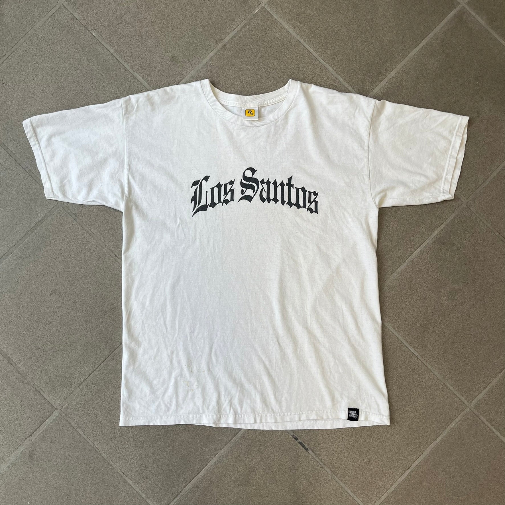 GTA V Los Santos 2013 White Tee features a white design with a large “Los Santos” graphic on the front in a tattoo-style font. The lower front includes a GTA V tag detail, while the neck tag features the Rockstar logo. The back has a small black Rockstar logo.
