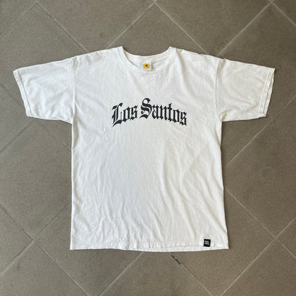 GTA V Los Santos 2013 White Tee features a white design with a large “Los Santos” graphic on the front in a tattoo-style font. The lower front includes a GTA V tag detail, while the neck tag features the Rockstar logo. The back has a small black Rockstar logo.
