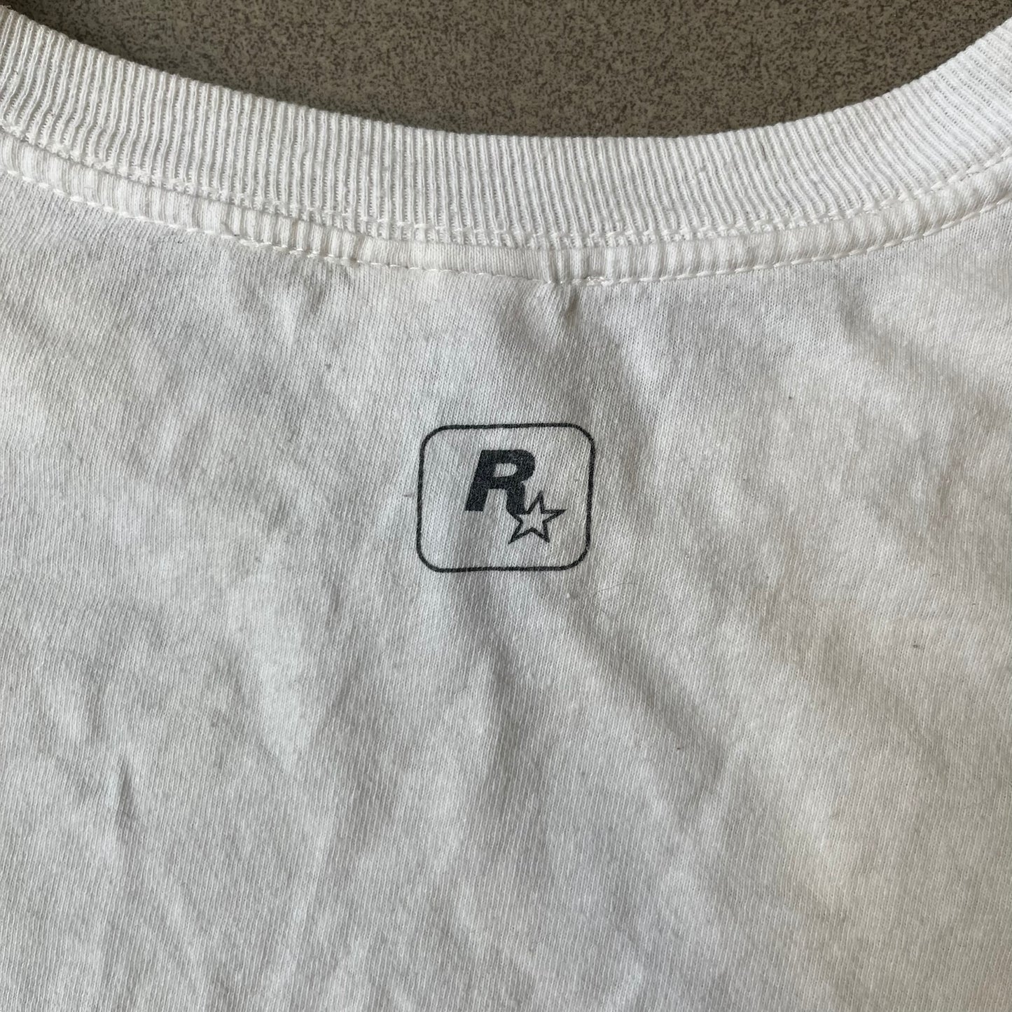 GTA V Los Santos 2013 White Tee features a white design with a large “Los Santos” graphic on the front in a tattoo-style font. The lower front includes a GTA V tag detail, while the neck tag features the Rockstar logo. The back has a small black Rockstar logo.
