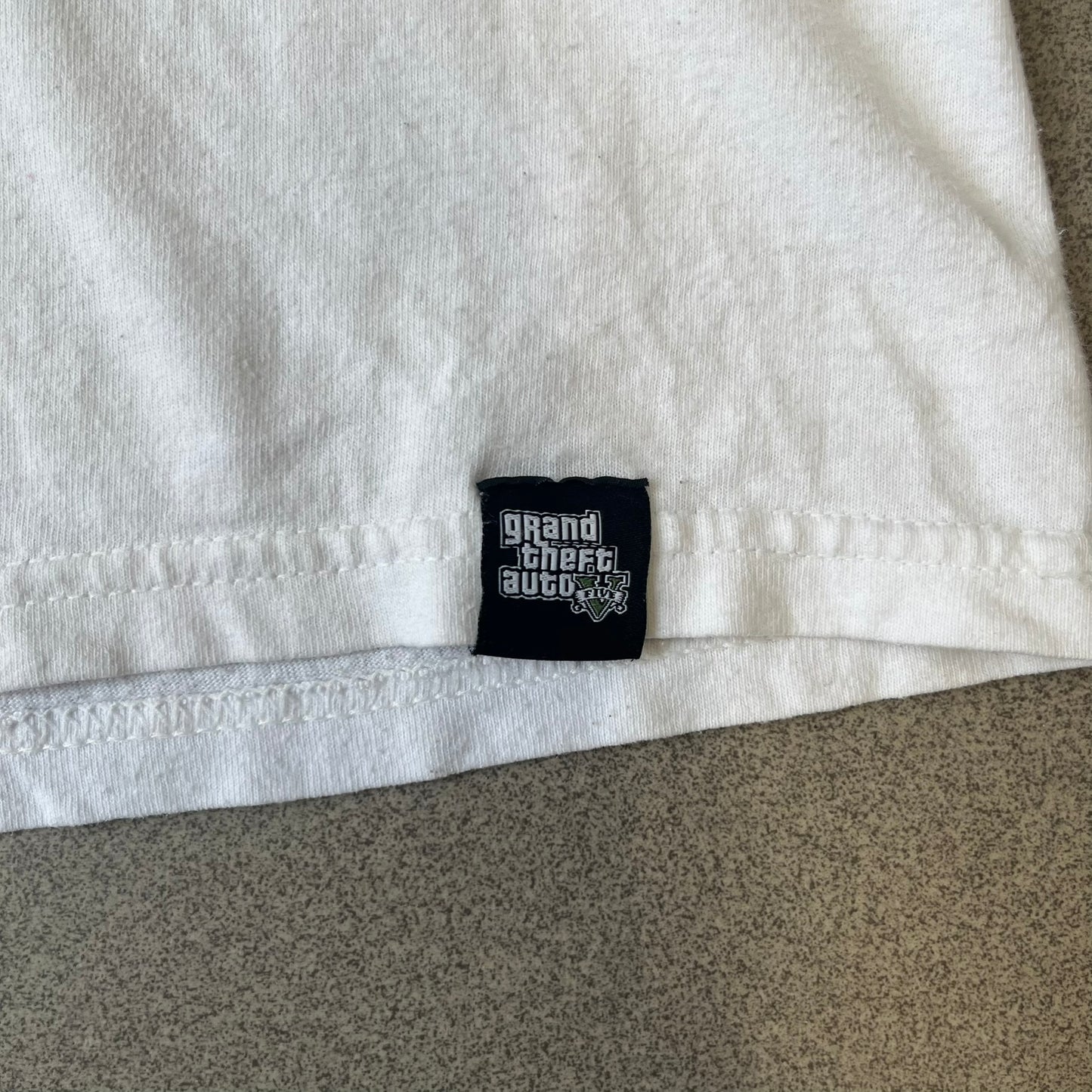 GTA V Los Santos 2013 White Tee features a white design with a large “Los Santos” graphic on the front in a tattoo-style font. The lower front includes a GTA V tag detail, while the neck tag features the Rockstar logo. The back has a small black Rockstar logo.

