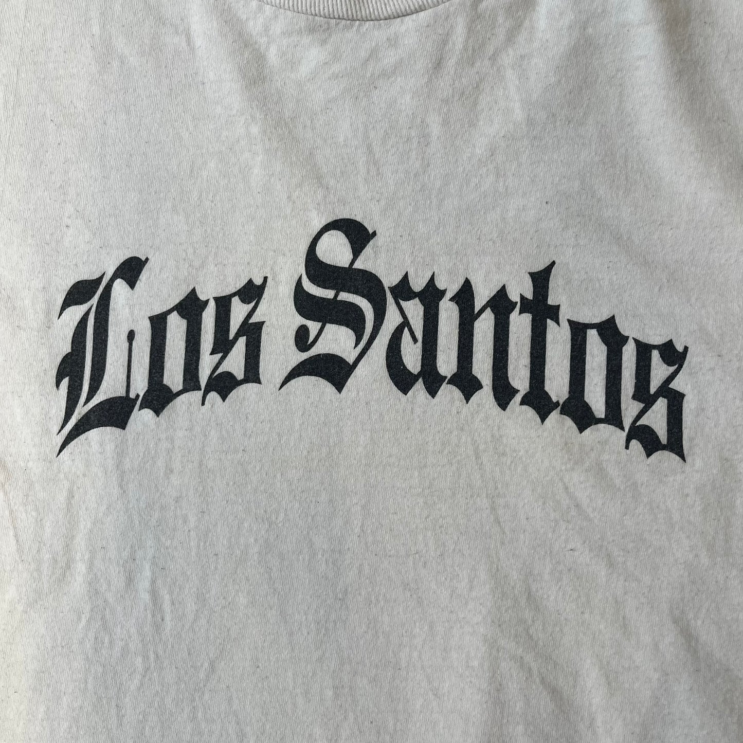 GTA V Los Santos 2013 White Tee features a white design with a large “Los Santos” graphic on the front in a tattoo-style font. The lower front includes a GTA V tag detail, while the neck tag features the Rockstar logo. The back has a small black Rockstar logo.
