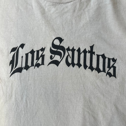 GTA V Los Santos 2013 White Tee features a white design with a large “Los Santos” graphic on the front in a tattoo-style font. The lower front includes a GTA V tag detail, while the neck tag features the Rockstar logo. The back has a small black Rockstar logo.
