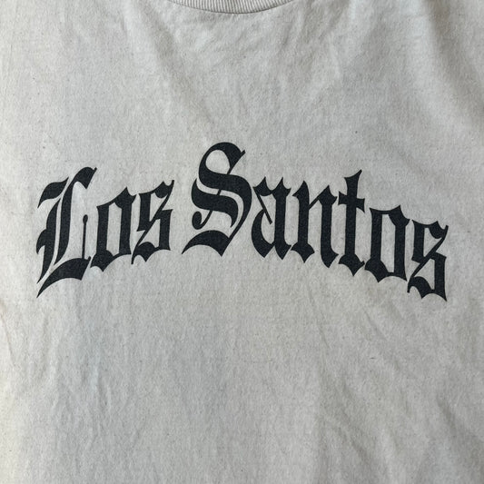 GTA V Los Santos 2013 White Tee features a white design with a large “Los Santos” graphic on the front in a tattoo-style font. The lower front includes a GTA V tag detail, while the neck tag features the Rockstar logo. The back has a small black Rockstar logo.
