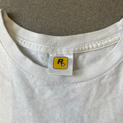 GTA V Los Santos 2013 White Tee features a white design with a large “Los Santos” graphic on the front in a tattoo-style font. The lower front includes a GTA V tag detail, while the neck tag features the Rockstar logo. The back has a small black Rockstar logo.
