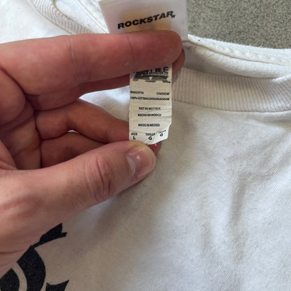 GTA V Los Santos 2013 White Tee features a white design with a large “Los Santos” graphic on the front in a tattoo-style font. The lower front includes a GTA V tag detail, while the neck tag features the Rockstar logo. The back has a small black Rockstar logo.
