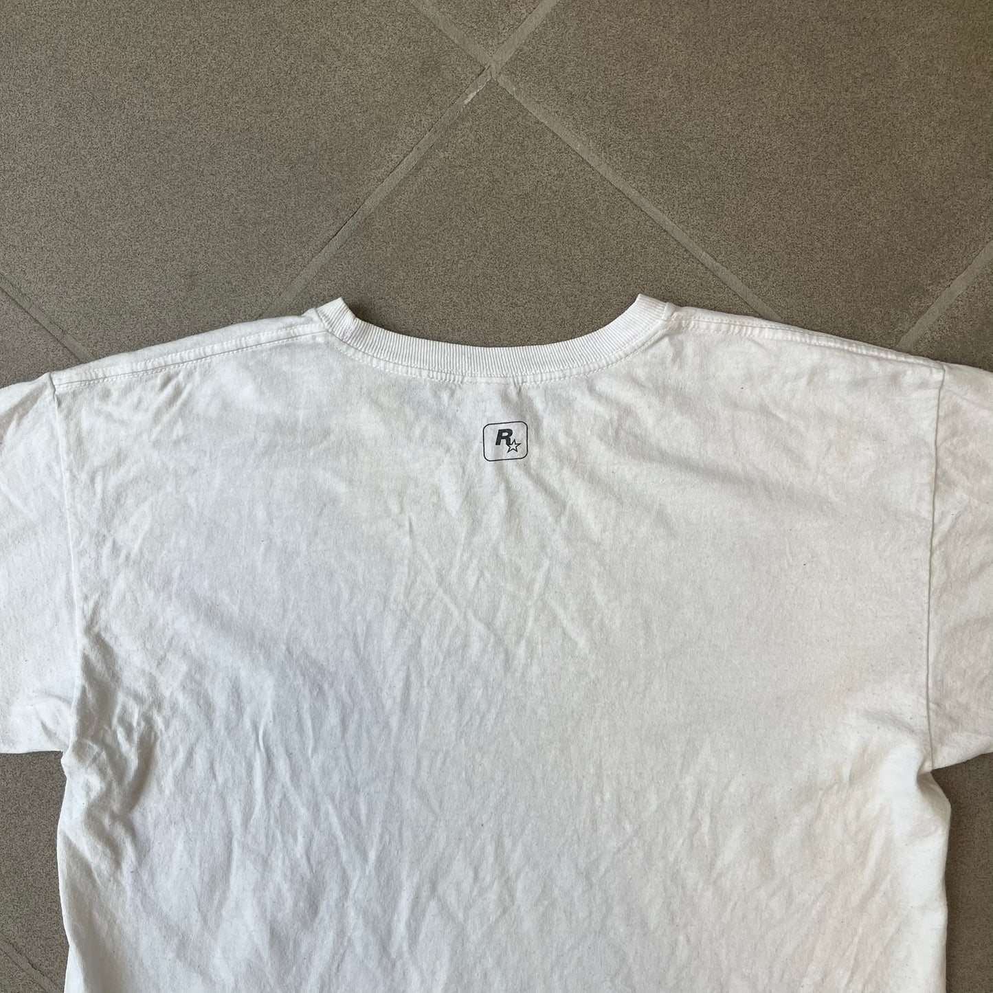 GTA V Los Santos 2013 White Tee features a white design with a large “Los Santos” graphic on the front in a tattoo-style font. The lower front includes a GTA V tag detail, while the neck tag features the Rockstar logo. The back has a small black Rockstar logo.
