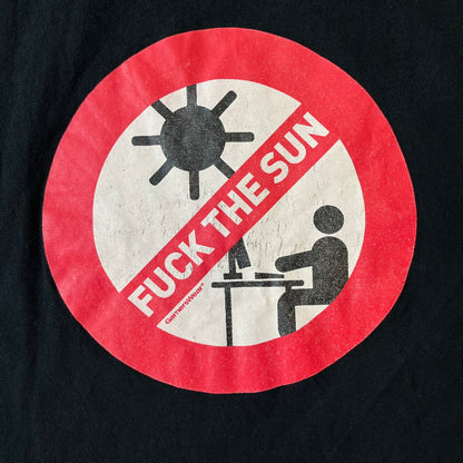 GamersWear 2010's Fuck The Sun Black Tee features a black 100% cotton T-shirt with a road sign-style graphic on the front. The graphic shows a black figure sitting at a computer beneath a black sun, surrounded by a white background and red outline. A red line crosses the design with white “Fuck The Sun” text, along with small red GamersWear branding.
