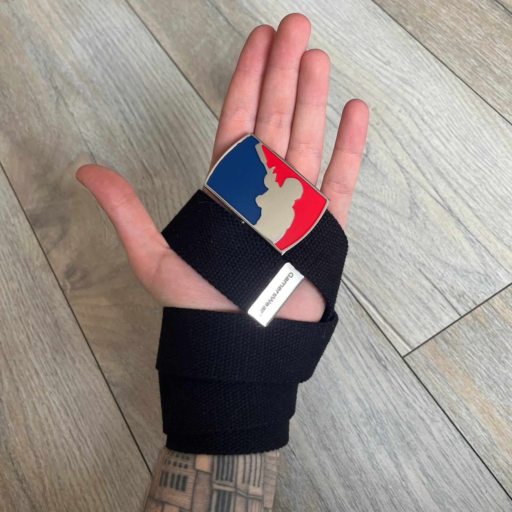 Gamerswear Counter-Strike 2016 Black Belt is a black cotton belt with an adjustable fit and a metal tip featuring a cutout Gamerswear logo. The buckle showcases an armed character in a silver finish, framed by blue enamel on the left side and red enamel on the right. The central figure is highlighted with a raised silver design and matching silver border detailing.