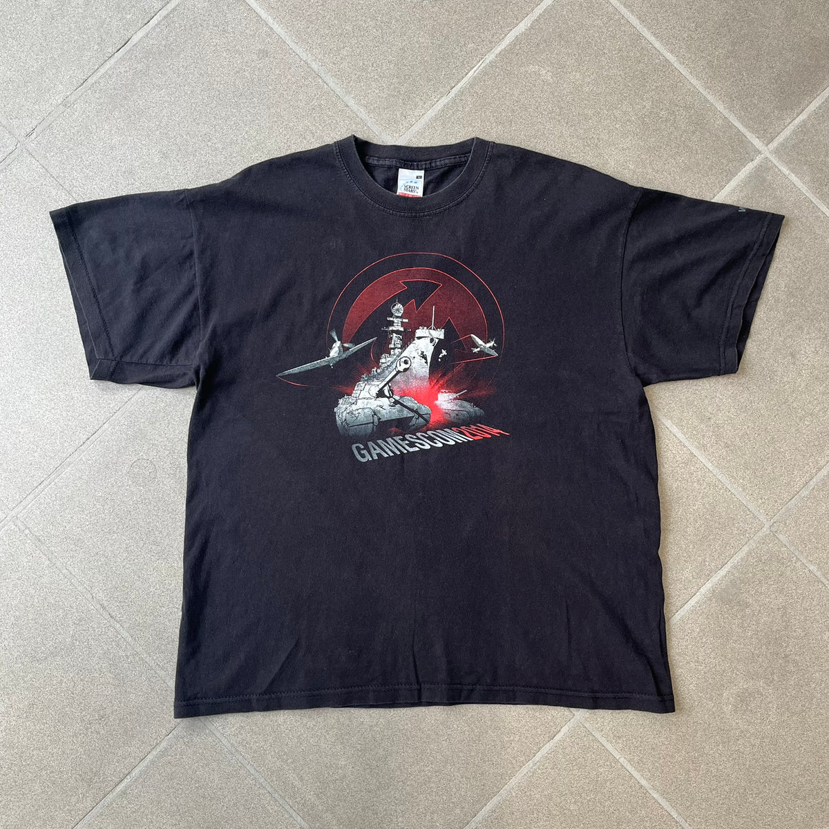 Gamescom 2014 WOT, Warplanes & Warships Promo Tee features a black 100% cotton T-shirt made on a Screen Stars blank. The front has a white and red graphic depicting a plane, tank, and warship, with “gamescom 2014” text underneath. The upper back has World of Tanks, World of Warplanes, and World of Warships logos, while the sleeve features a Wargaming.net logo.
