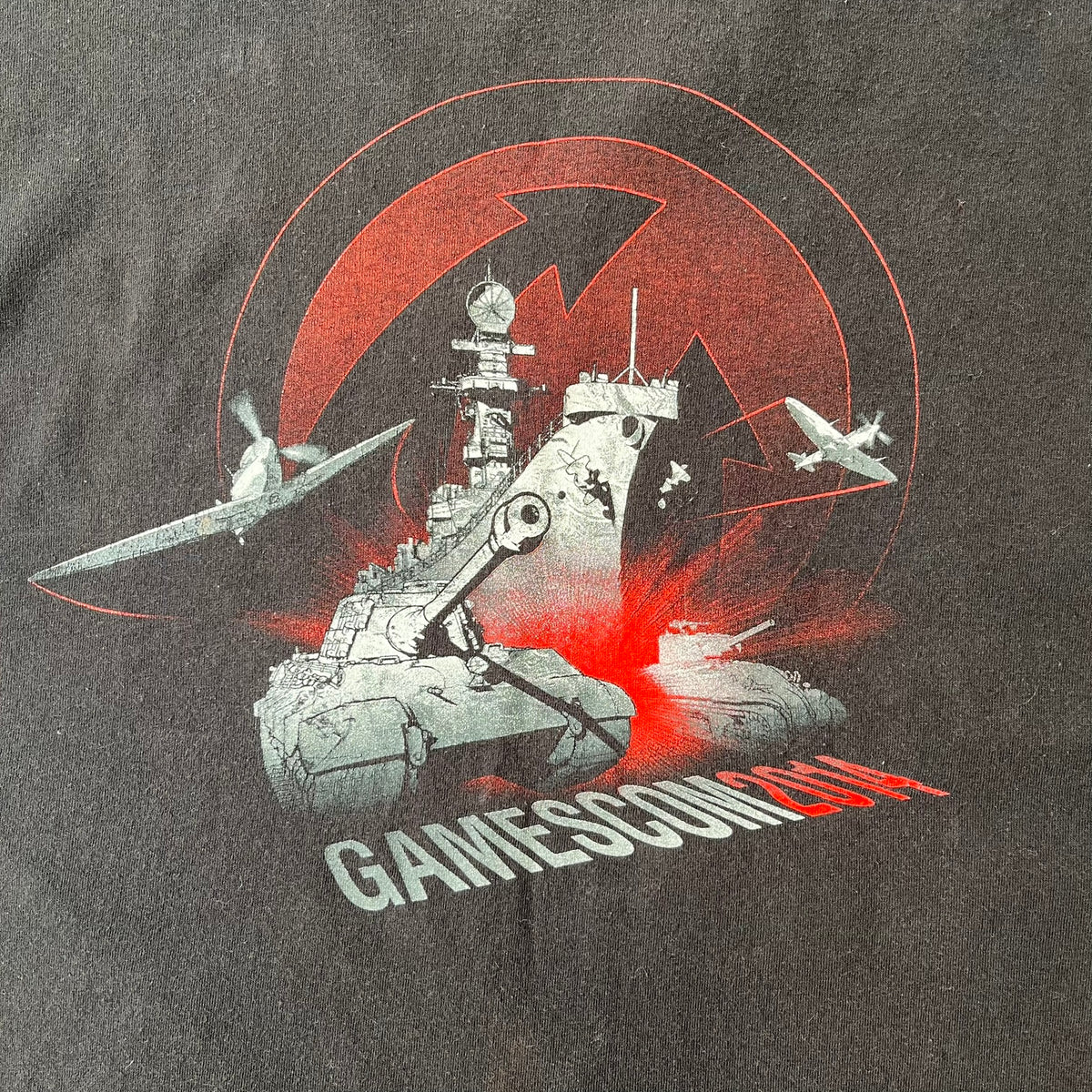 Gamescom 2014 WOT, Warplanes & Warships Promo Tee features a black 100% cotton T-shirt made on a Screen Stars blank. The front has a white and red graphic depicting a plane, tank, and warship, with “gamescom 2014” text underneath. The upper back has World of Tanks, World of Warplanes, and World of Warships logos, while the sleeve features a Wargaming.net logo.

