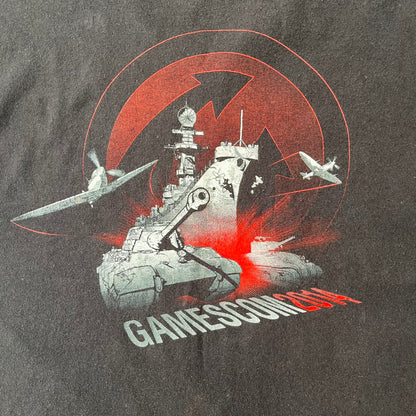 Gamescom 2014 WOT, Warplanes & Warships Promo Tee features a black 100% cotton T-shirt made on a Screen Stars blank. The front has a white and red graphic depicting a plane, tank, and warship, with “gamescom 2014” text underneath. The upper back has World of Tanks, World of Warplanes, and World of Warships logos, while the sleeve features a Wargaming.net logo.
