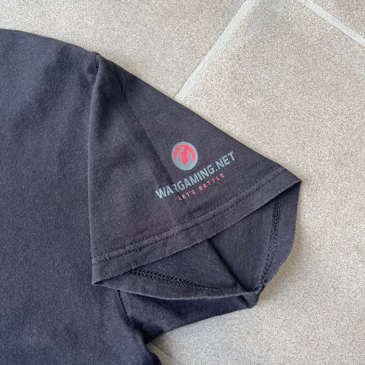 Gamescom 2014 WOT, Warplanes & Warships Promo Tee features a black 100% cotton T-shirt made on a Screen Stars blank. The front has a white and red graphic depicting a plane, tank, and warship, with “gamescom 2014” text underneath. The upper back has World of Tanks, World of Warplanes, and World of Warships logos, while the sleeve features a Wargaming.net logo.
