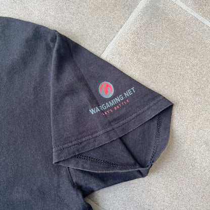 Gamescom 2014 WOT, Warplanes & Warships Promo Tee features a black 100% cotton T-shirt made on a Screen Stars blank. The front has a white and red graphic depicting a plane, tank, and warship, with “gamescom 2014” text underneath. The upper back has World of Tanks, World of Warplanes, and World of Warships logos, while the sleeve features a Wargaming.net logo.

