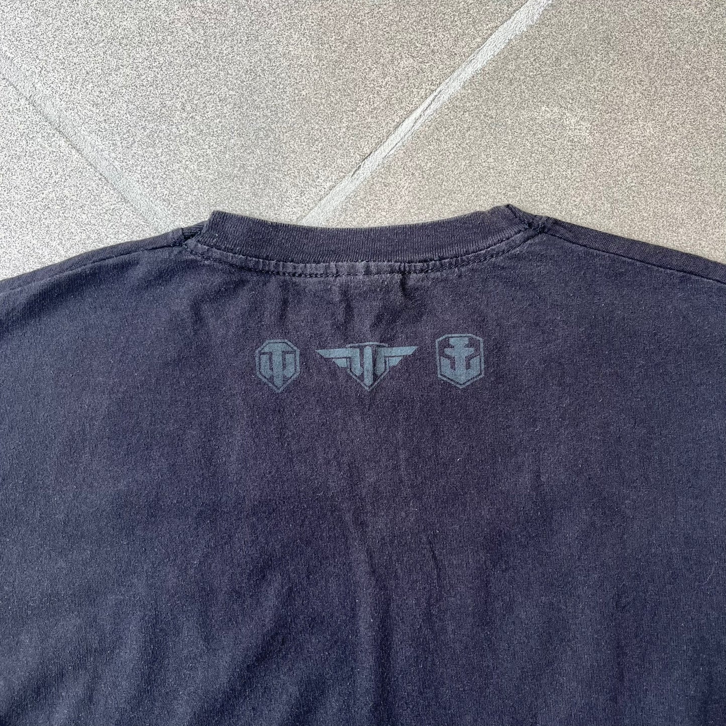 Gamescom 2014 WOT, Warplanes & Warships Promo Tee features a black 100% cotton T-shirt made on a Screen Stars blank. The front has a white and red graphic depicting a plane, tank, and warship, with “gamescom 2014” text underneath. The upper back has World of Tanks, World of Warplanes, and World of Warships logos, while the sleeve features a Wargaming.net logo.
