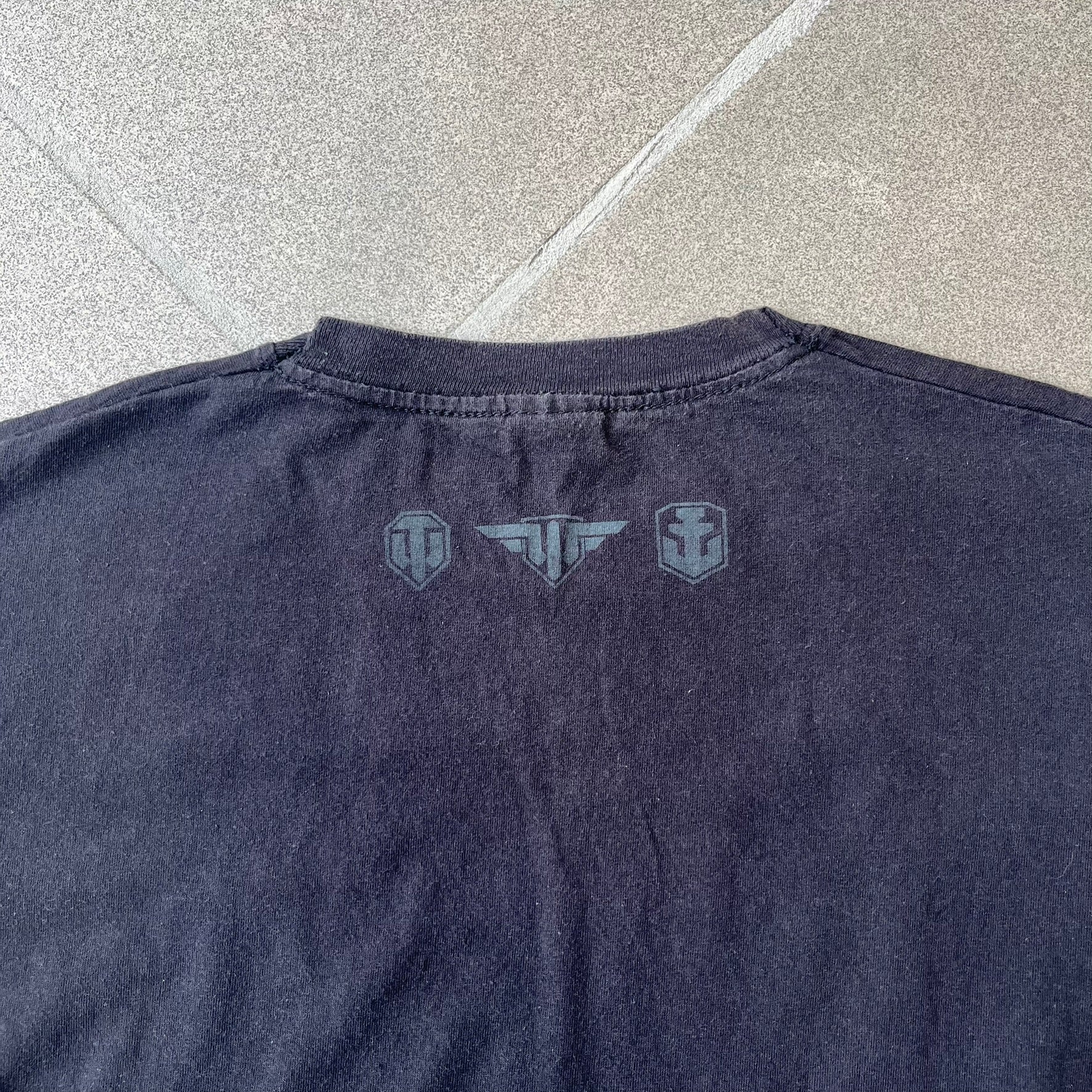 Gamescom 2014 WOT, Warplanes & Warships Promo Tee features a black 100% cotton T-shirt made on a Screen Stars blank. The front has a white and red graphic depicting a plane, tank, and warship, with “gamescom 2014” text underneath. The upper back has World of Tanks, World of Warplanes, and World of Warships logos, while the sleeve features a Wargaming.net logo.
