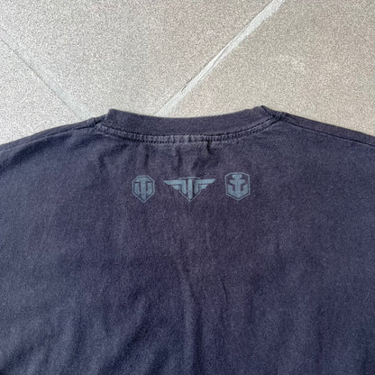 Gamescom 2014 WOT, Warplanes & Warships Promo Tee features a black 100% cotton T-shirt made on a Screen Stars blank. The front has a white and red graphic depicting a plane, tank, and warship, with “gamescom 2014” text underneath. The upper back has World of Tanks, World of Warplanes, and World of Warships logos, while the sleeve features a Wargaming.net logo.
