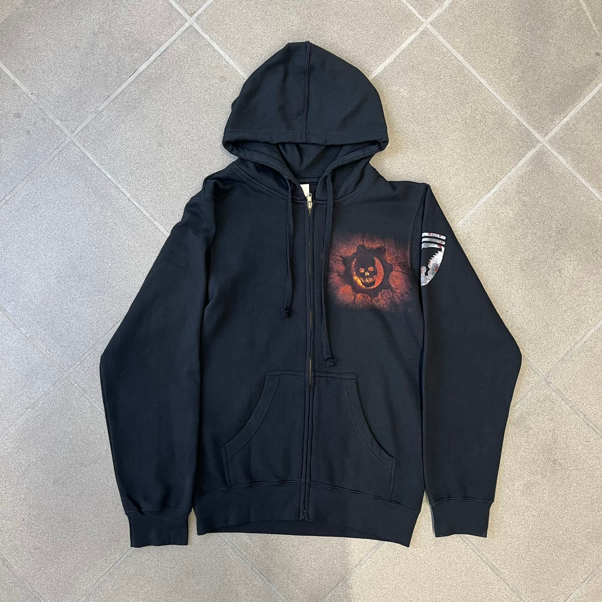 Gears of War 3 Marcus Fenix 2010 Promo Hoodie is a black zip-up hoodie with a dark Crimson Omen logo in orange tones on the left upper chest and a bloody Delta-One logo on one sleeve. The back features a large Marcus Fenix graphic with “Gears of War 3” text below it and a small 2010 Epic Games licensing mark.
