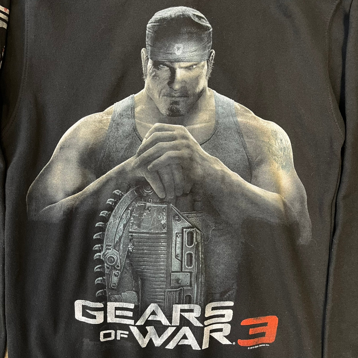 Gears of War 3 Marcus Fenix 2010 Promo Hoodie is a black zip-up hoodie with a dark Crimson Omen logo in orange tones on the left upper chest and a bloody Delta-One logo on one sleeve. The back features a large Marcus Fenix graphic with “Gears of War 3” text below it and a small 2010 Epic Games licensing mark.