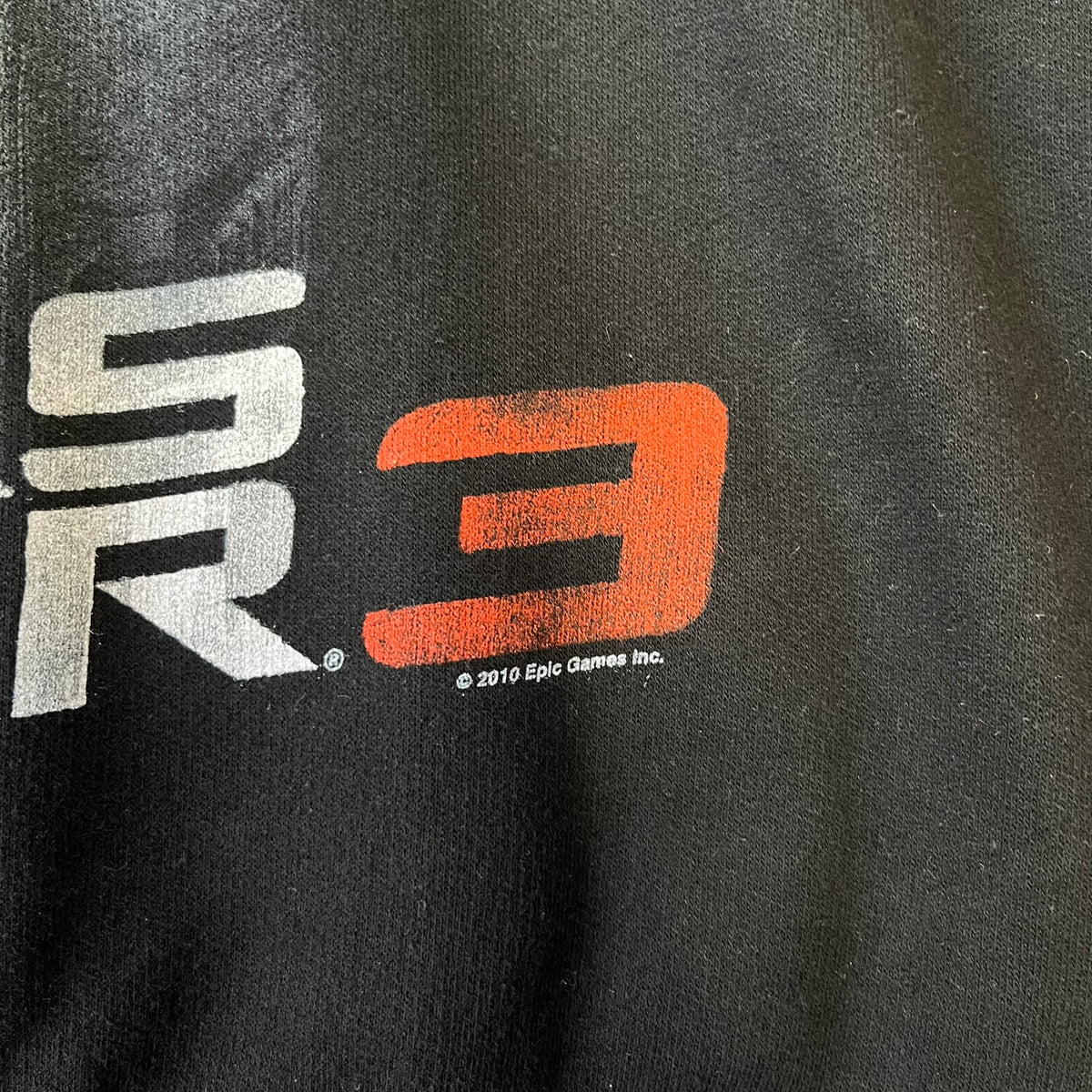 Gears of War 3 Marcus Fenix 2010 Promo Hoodie is a black zip-up hoodie with a dark Crimson Omen logo in orange tones on the left upper chest and a bloody Delta-One logo on one sleeve. The back features a large Marcus Fenix graphic with “Gears of War 3” text below it and a small 2010 Epic Games licensing mark.