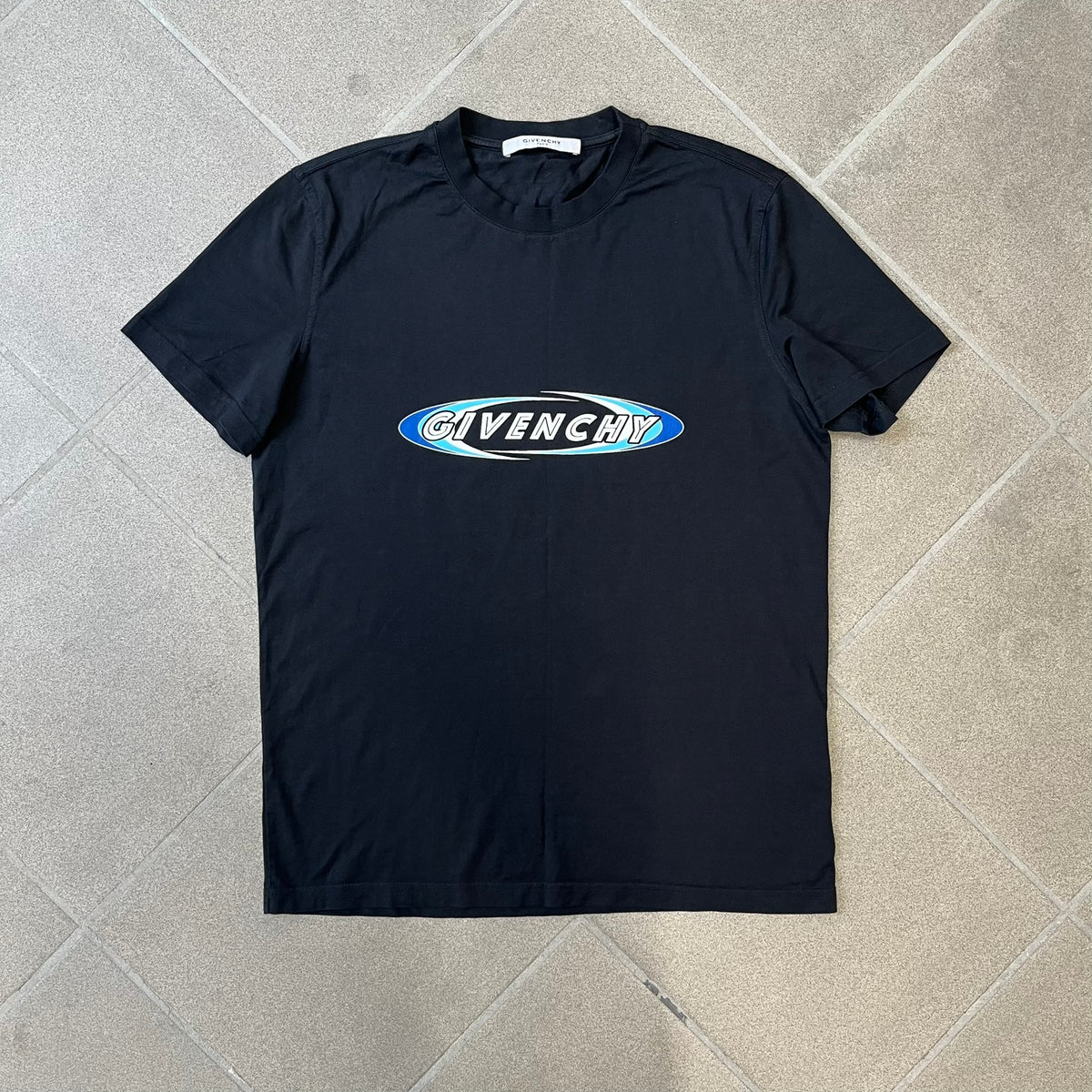 Givenchy AW18 Aquarius Black Tee is a black, slim-fit T-shirt made from 100% cotton, featuring white "GIVENCHY" text on the front alongside a blue half-moon-shaped graphics. The back displays an Aquarius-inspired design in white, gray, and blue, depicting a fountain with a large bird, a cube built from repeated Givenchy logos, and a Givenchy signature.
