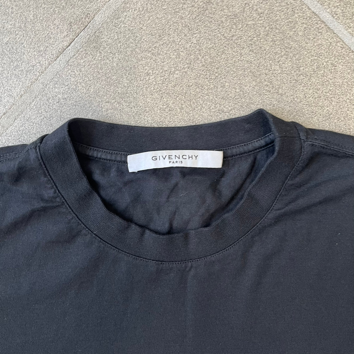 Givenchy AW18 Aquarius Black Tee is a black, slim-fit T-shirt made from 100% cotton, featuring white "GIVENCHY" text on the front alongside a blue half-moon-shaped graphics. The back displays an Aquarius-inspired design in white, gray, and blue, depicting a fountain with a large bird, a cube built from repeated Givenchy logos, and a Givenchy signature.