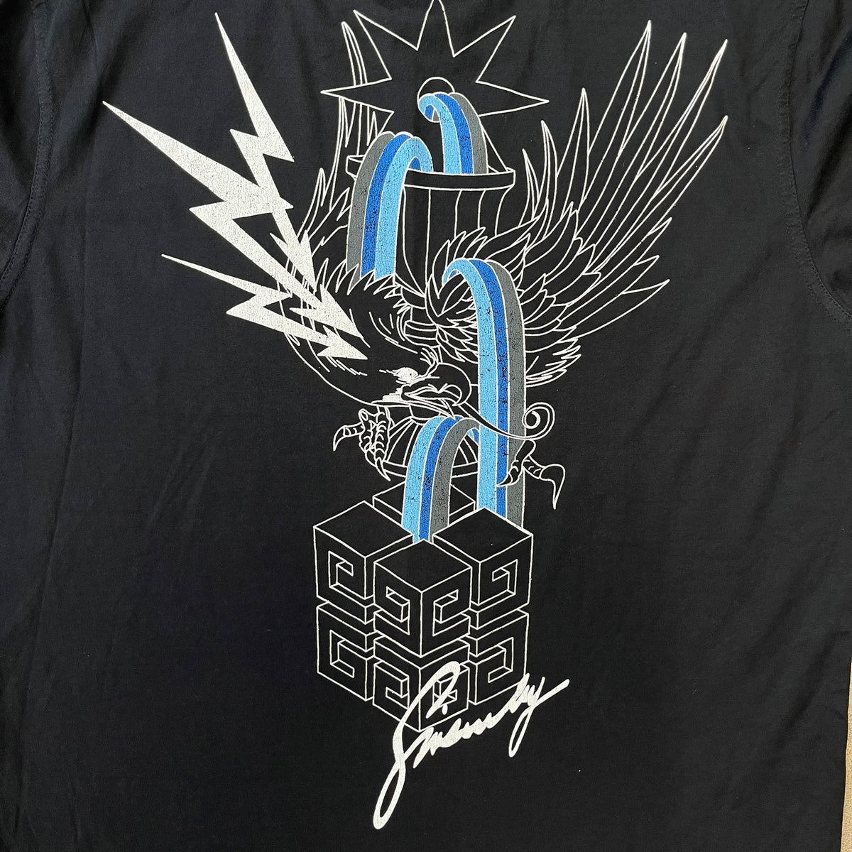 Givenchy AW18 Aquarius Black Tee is a black, slim-fit T-shirt made from 100% cotton, featuring white "GIVENCHY" text on the front alongside a blue half-moon-shaped graphics. The back displays an Aquarius-inspired design in white, gray, and blue, depicting a fountain with a large bird, a cube built from repeated Givenchy logos, and a Givenchy signature.