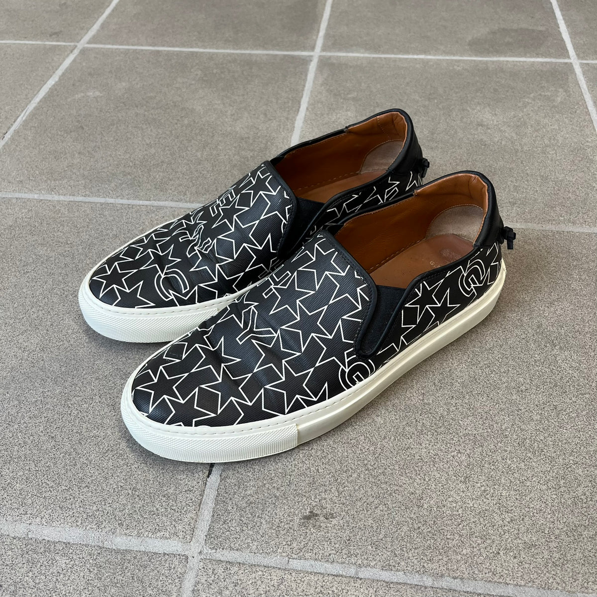 Givenchy Black & White Stars Slip-On Sneakers are made in Italy from premium black leather, featuring a white sole and a black upper detailed with a white outlined pattern of Givenchy text and stars. The interior is finished in brown, with a small Givenchy logo placed on the back of the sole.