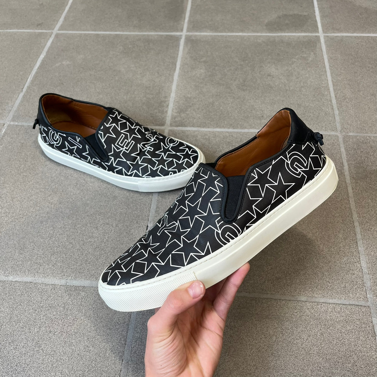 Givenchy Black & White Stars Slip-On Sneakers are made in Italy from premium black leather, featuring a white sole and a black upper detailed with a white outlined pattern of Givenchy text and stars. The interior is finished in brown, with a small Givenchy logo placed on the back of the sole.