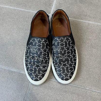 Givenchy Black & White Stars Slip-On Sneakers are made in Italy from premium black leather, featuring a white sole and a black upper detailed with a white outlined pattern of Givenchy text and stars. The interior is finished in brown, with a small Givenchy logo placed on the back of the sole.