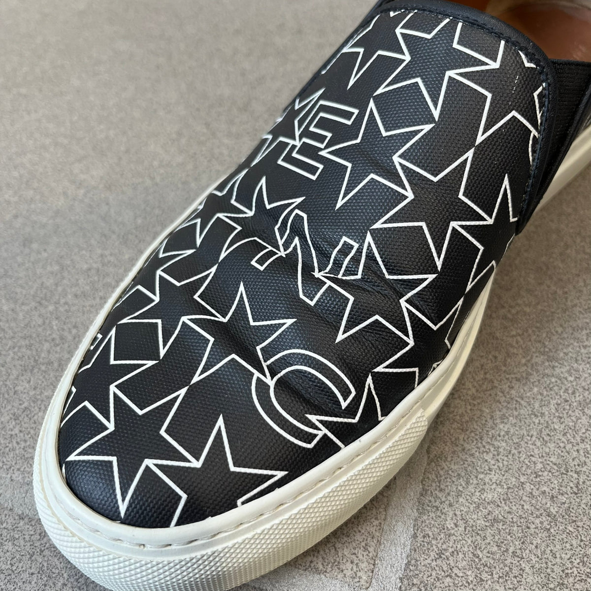 Givenchy Black & White Stars Slip-On Sneakers are made in Italy from premium black leather, featuring a white sole and a black upper detailed with a white outlined pattern of Givenchy text and stars. The interior is finished in brown, with a small Givenchy logo placed on the back of the sole.