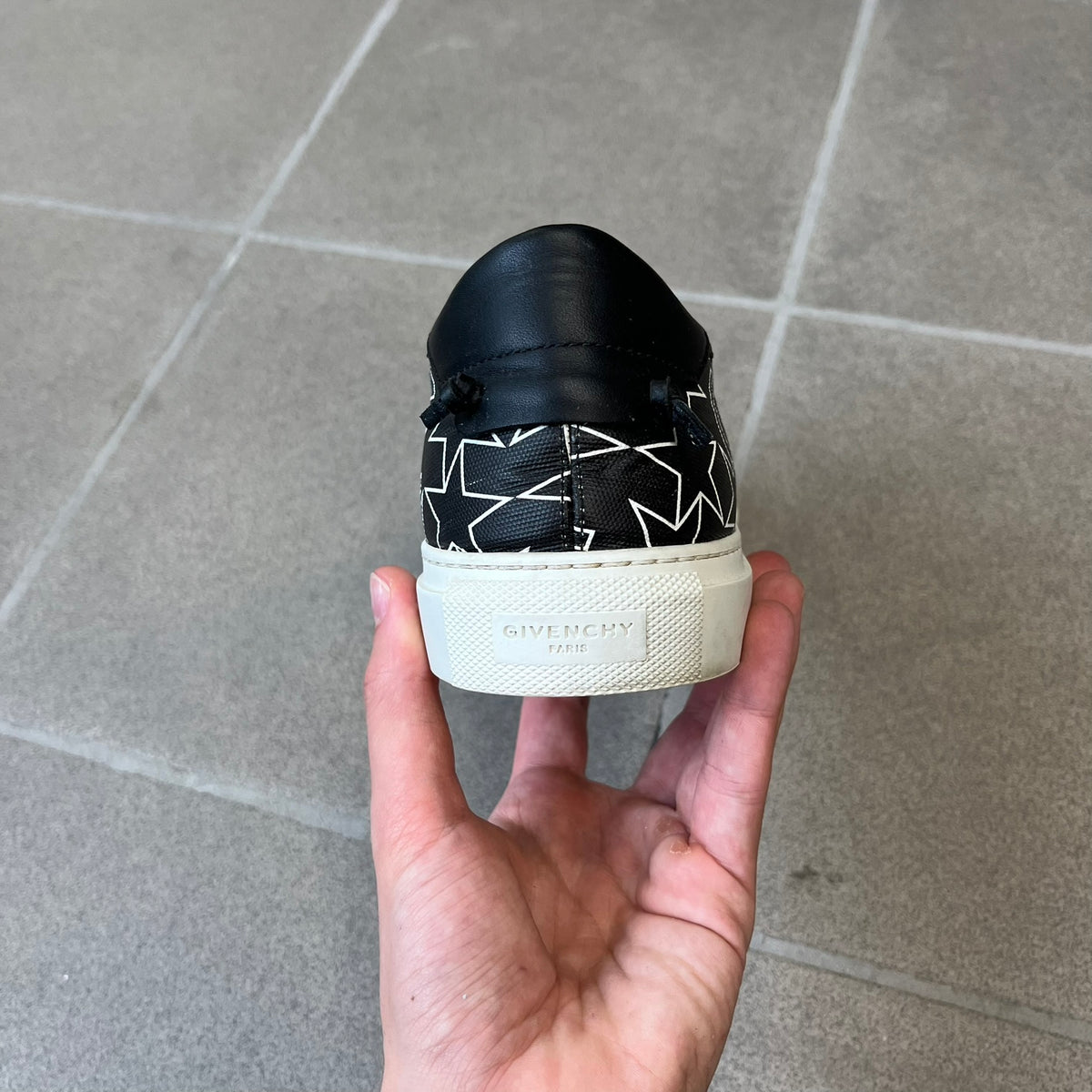 Givenchy Black & White Stars Slip-On Sneakers are made in Italy from premium black leather, featuring a white sole and a black upper detailed with a white outlined pattern of Givenchy text and stars. The interior is finished in brown, with a small Givenchy logo placed on the back of the sole.
