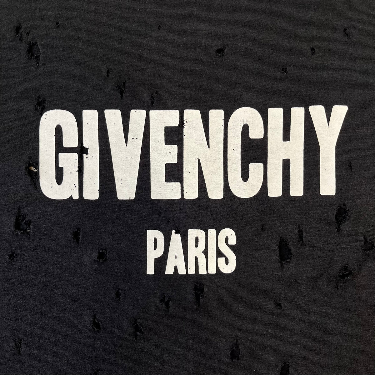 Givenchy Distressed Logo Paris Tee is a black T-shirt made from 100% cotton in Portugal, featuring white "GIVENCHY" text on the chest with "PARIS" printed below it. The shirt is finished with an all-over factory-distressed design, with holes intentionally placed across both the front and back.