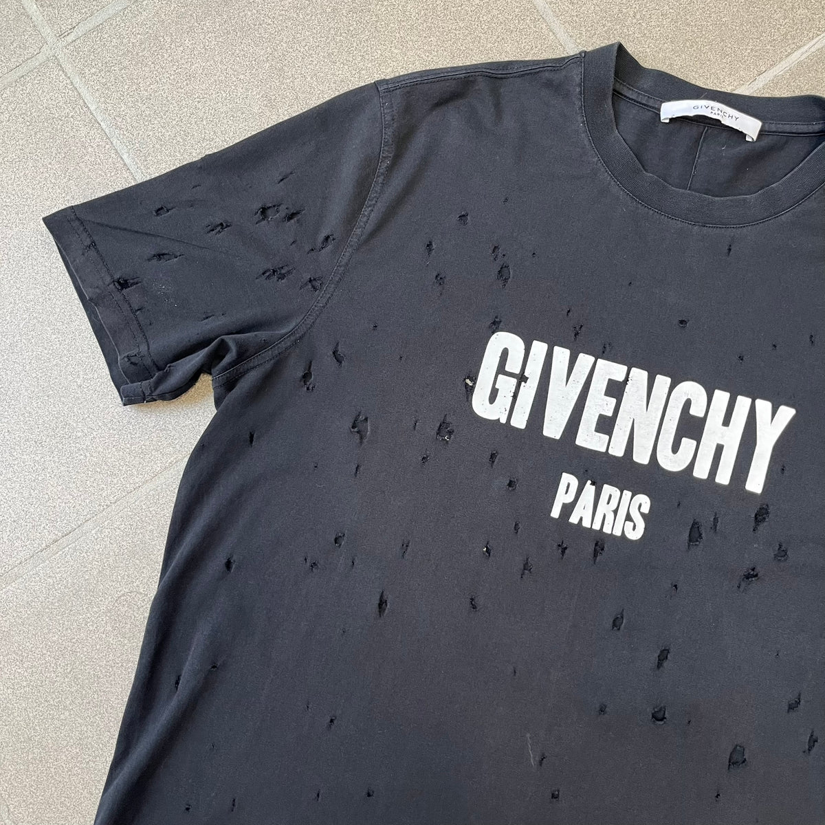 Givenchy Distressed Logo Paris Tee is a black T-shirt made from 100% cotton in Portugal, featuring white "GIVENCHY" text on the chest with "PARIS" printed below it. The shirt is finished with an all-over factory-distressed design, with holes intentionally placed across both the front and back.