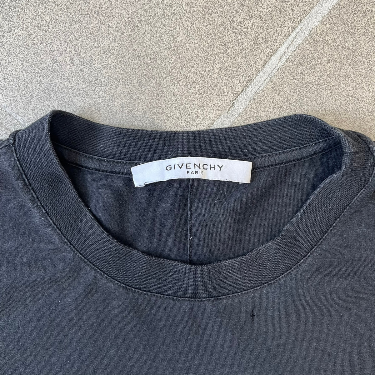 Givenchy Distressed Logo Paris Tee is a black T-shirt made from 100% cotton in Portugal, featuring white "GIVENCHY" text on the chest with "PARIS" printed below it. The shirt is finished with an all-over factory-distressed design, with holes intentionally placed across both the front and back.