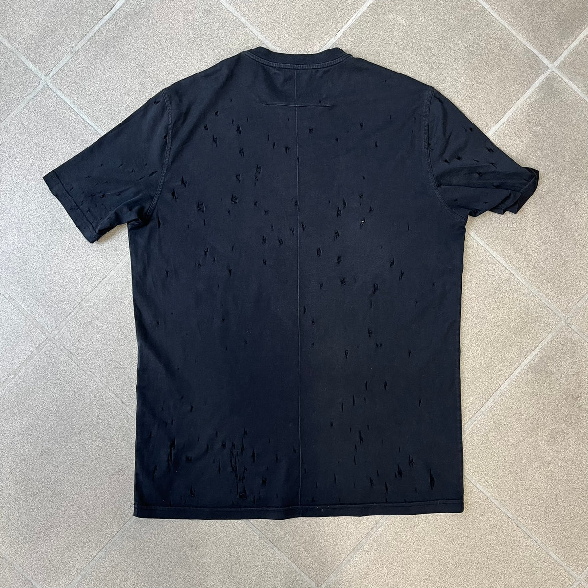 Givenchy Distressed Logo Paris Tee is a black T-shirt made from 100% cotton in Portugal, featuring white "GIVENCHY" text on the chest with "PARIS" printed below it. The shirt is finished with an all-over factory-distressed design, with holes intentionally placed across both the front and back.
