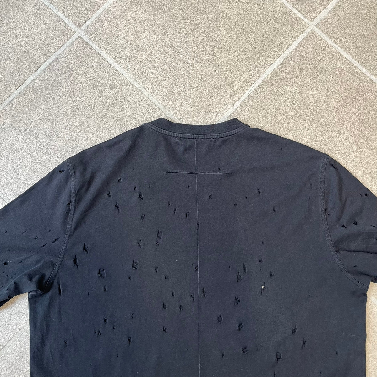 Givenchy Distressed Logo Paris Tee is a black T-shirt made from 100% cotton in Portugal, featuring white "GIVENCHY" text on the chest with "PARIS" printed below it. The shirt is finished with an all-over factory-distressed design, with holes intentionally placed across both the front and back.