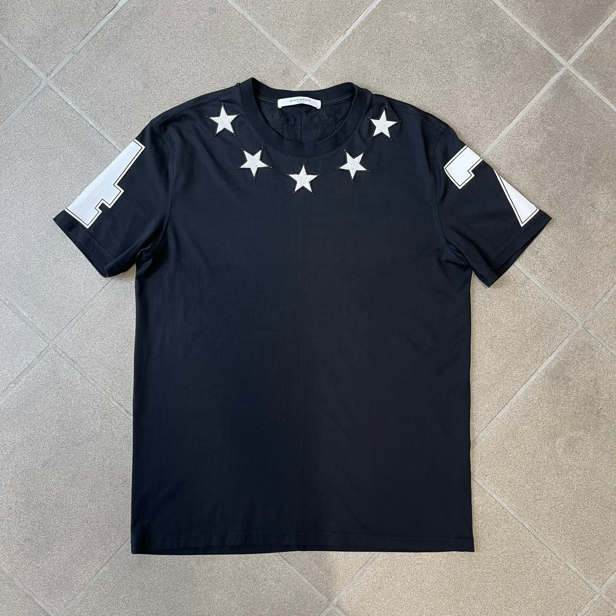 Givenchy 2017 Black Star Patch 4-7 Tee is a black, Cuban-fit T-shirt made from 100% cotton, featuring fuzzy white star patches trimming the collar on both the front and back. White "4" and "7" numbering is printed on the sleeves.
